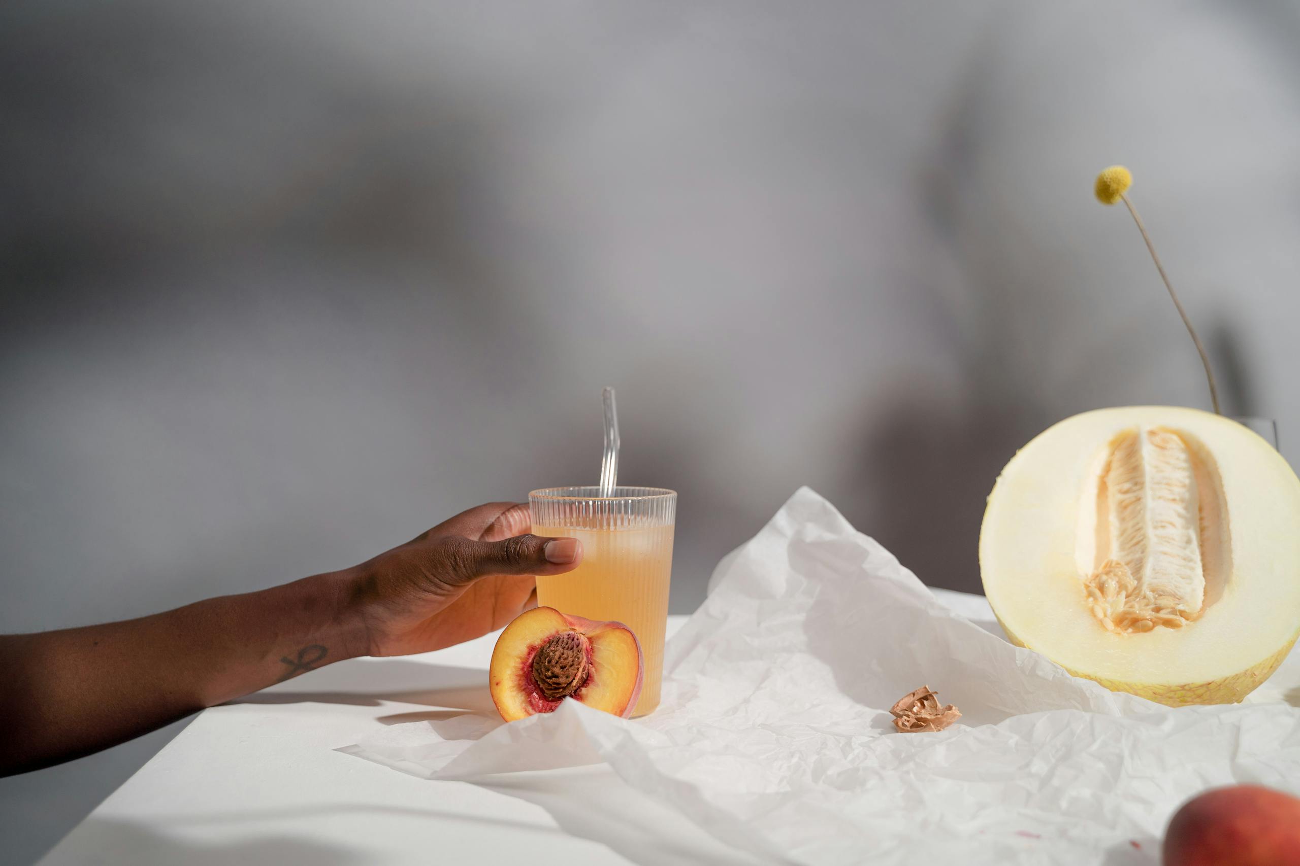 A refreshing glass of melon and peach juice being held by hand with fresh fruits and paper background.