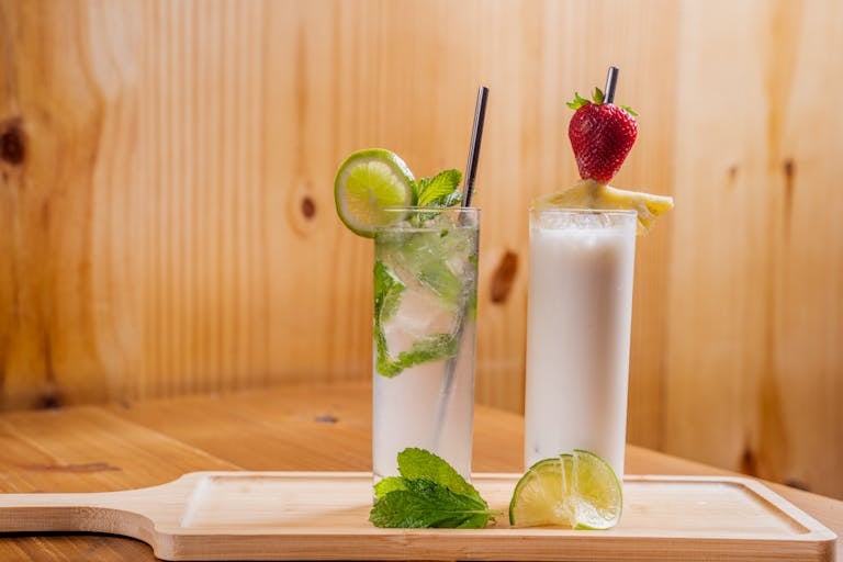 Two refreshing cocktails featuring lime, mint, and strawberry on a wooden board.