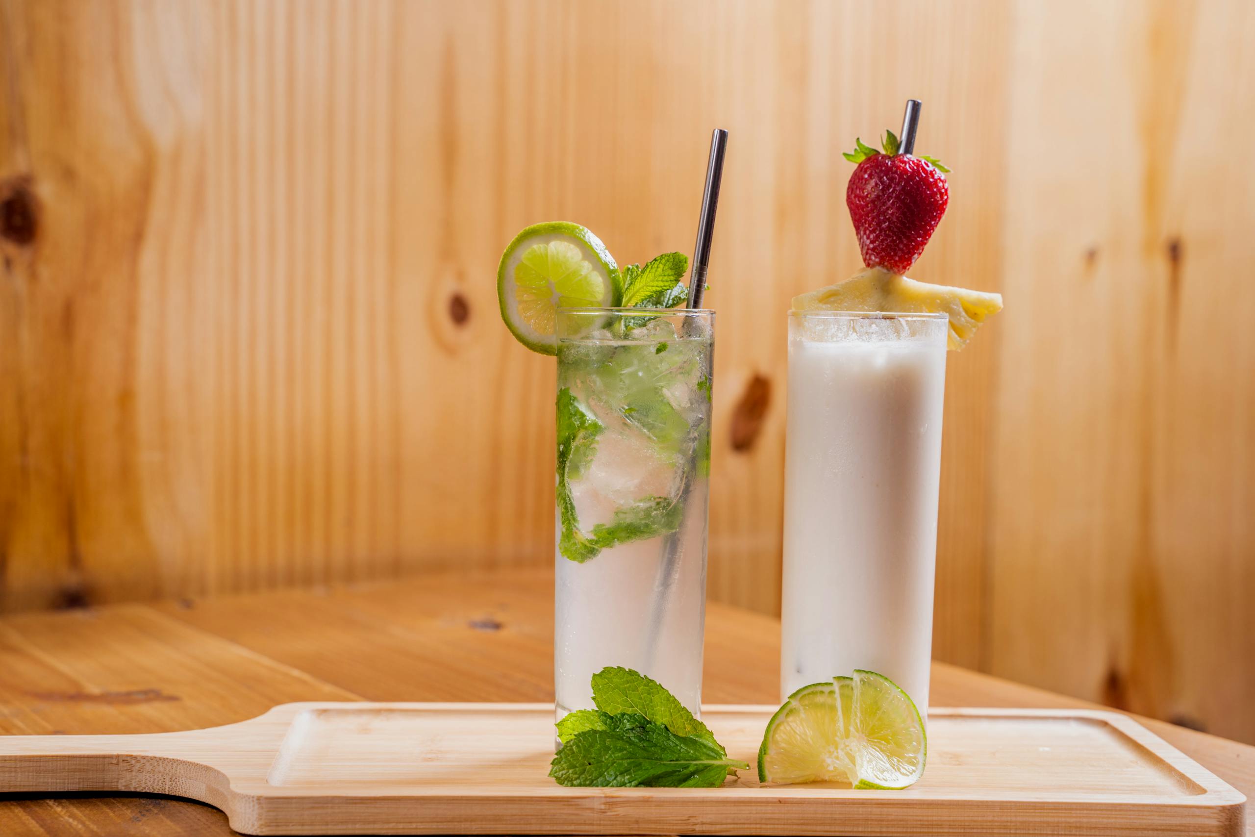Two refreshing cocktails featuring lime, mint, and strawberry on a wooden board.