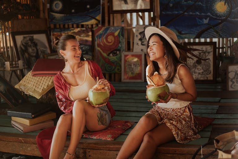 Two women laughing while enjoying refreshing coconut drinks in an artistic indoor setting.